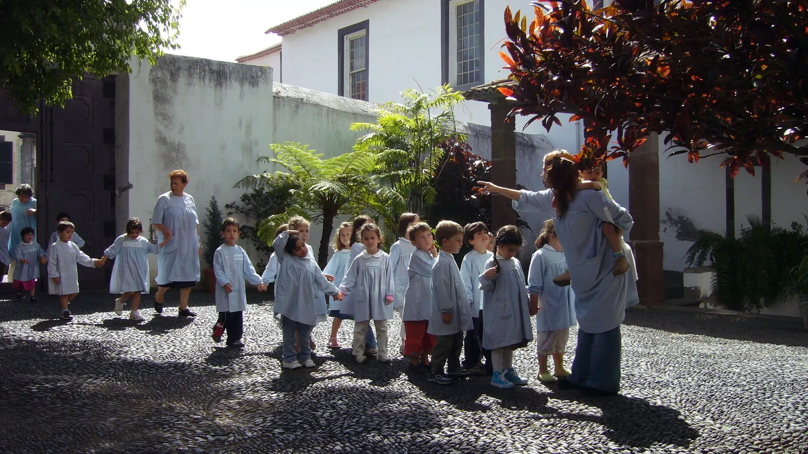 Kindergarten children in matching smocks walk hand in hand with their teachers across a cobbled courtyard in Funchal, Madeira, the kind of group outing typical of daycare in Portugal