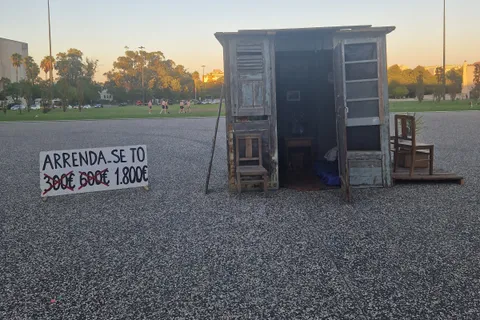 A plywood shack installed as a protest artwork outside the University of Lisbon, with a hand-painted sign reading Arrenda-se T0 and the rent crossed out from 300 to 600 to 1,800 euros, a comment on how hard renting in Portugal has become