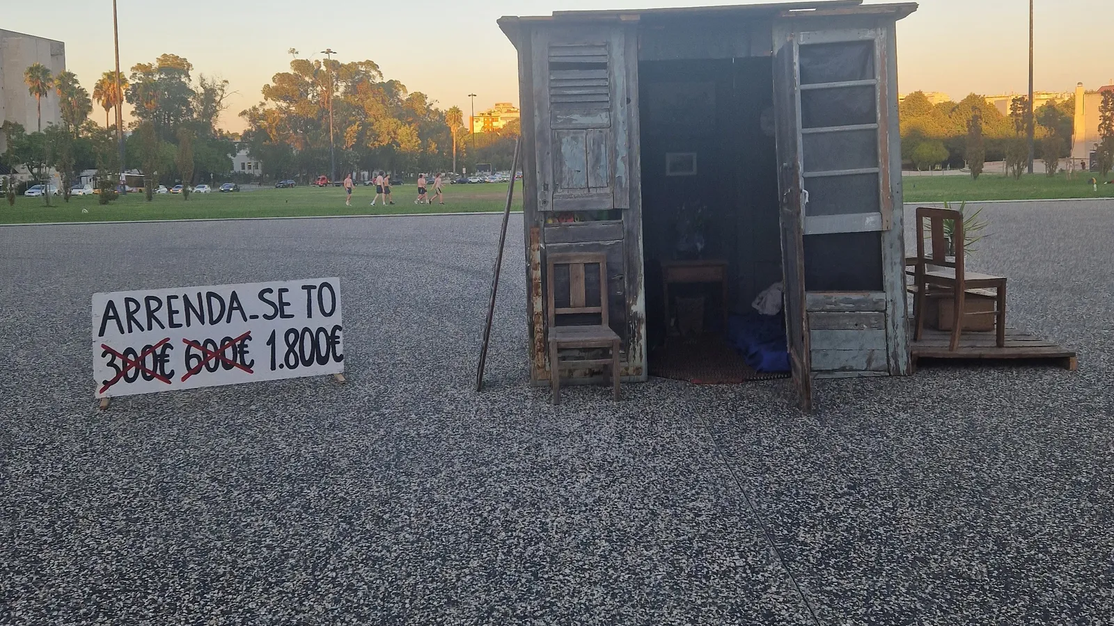 A plywood shack installed as a protest artwork outside the University of Lisbon, with a hand-painted sign reading Arrenda-se T0 and the rent crossed out from 300 to 600 to 1,800 euros, a comment on how hard renting in Portugal has become