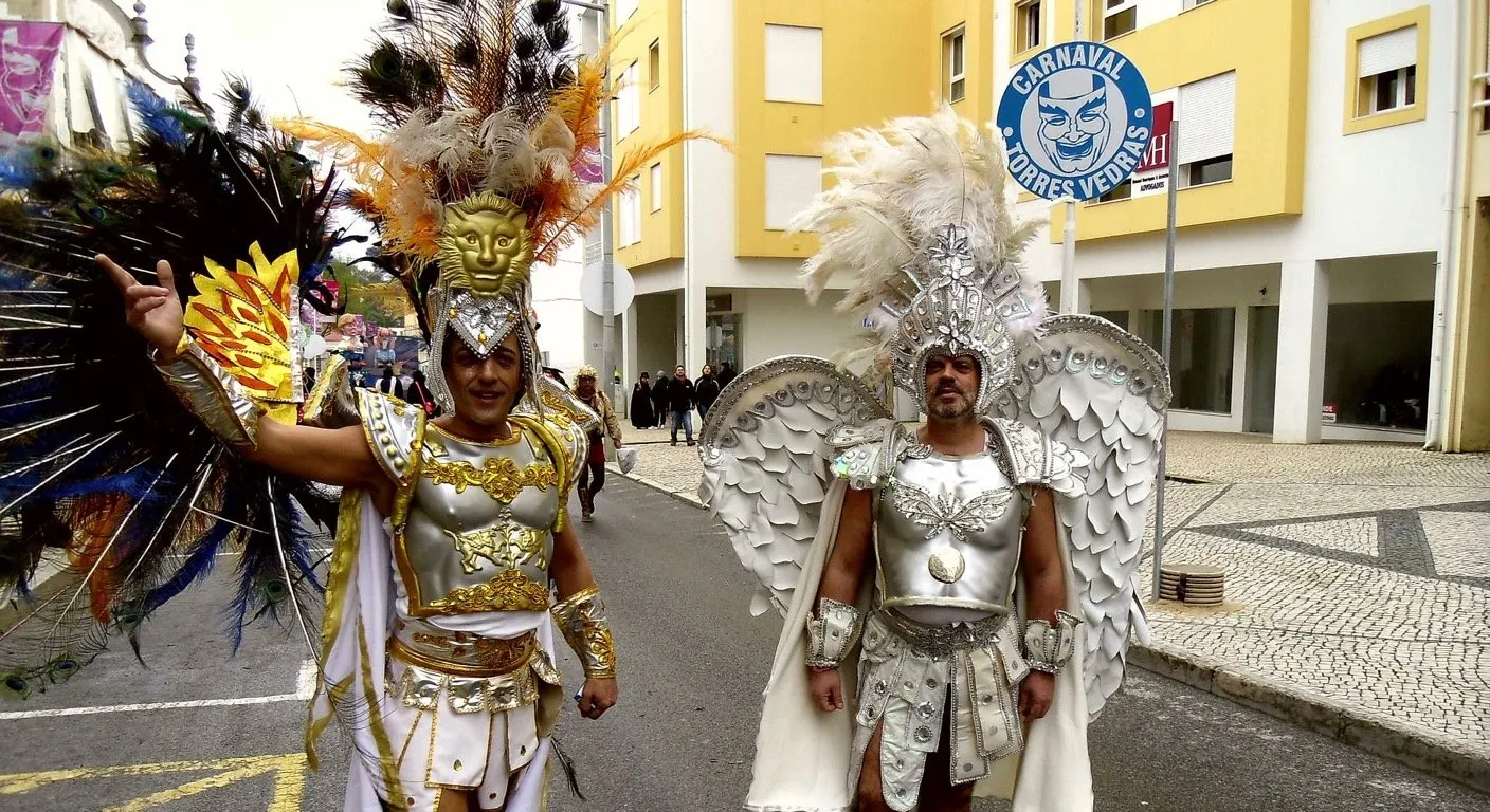 Costumed performers in feathered headdresses parading past a Carnaval de Torres Vedras sign in Portugal in February
