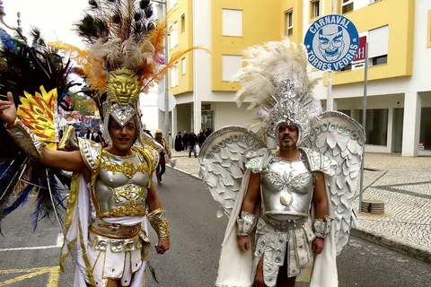 Costumed performers in feathered headdresses parading past a Carnaval de Torres Vedras sign in Portugal in February